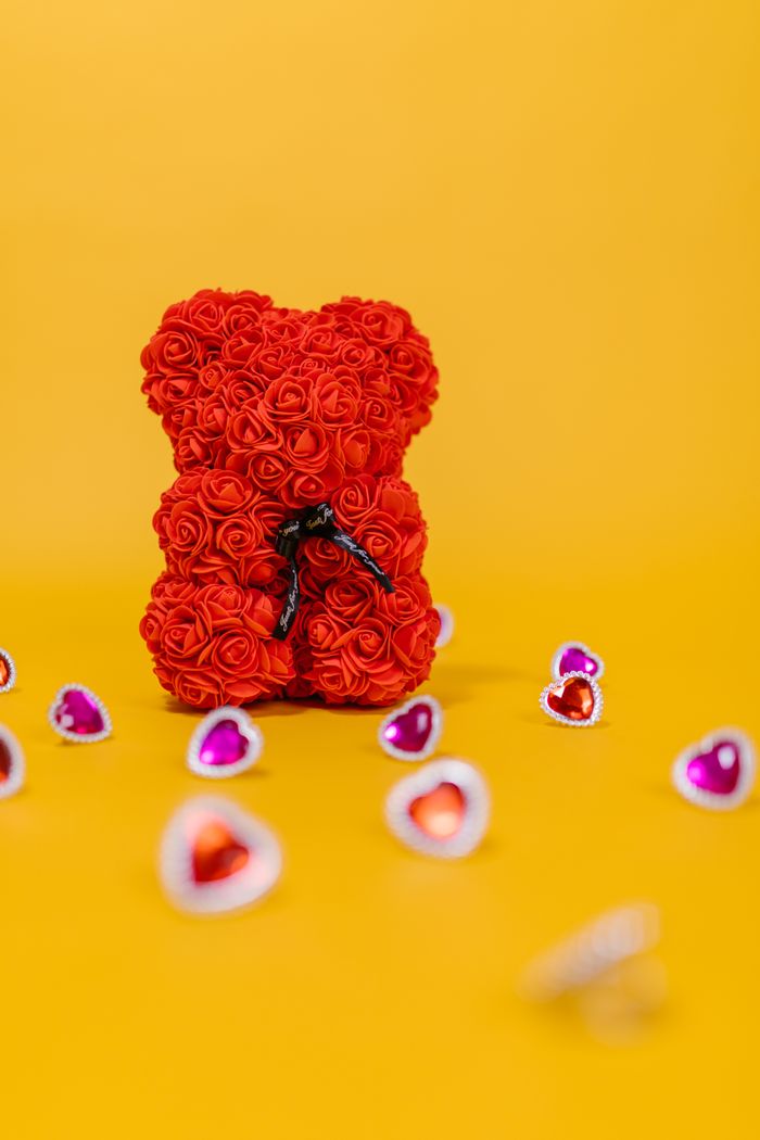 Red rose teddy bear surrounded by heart-shaped decorations on a yellow background, perfect for Valentine's Day.